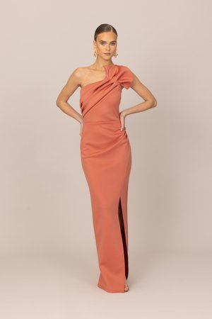 Kobieta w długiej do ziemi, koralowej sukience na jedno ramię z dużą falbaną i bocznym rozcięciem, pozująca z rękami na biodrach na jednolitym tle.