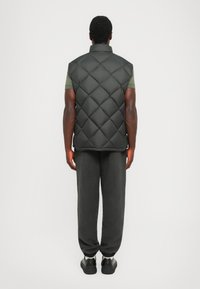 Vest puff cu model cu diamante, de culoare neagră, design fără mâneci, purtat peste o bluză verde. Asortat cu pantaloni închiși la culoare și adidași negri.