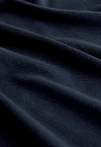 Jūras zila auduma ar gludu tekstūru, kurā redzami maigi viļņi un subtīls spīdums, izceļot materiāla smalko austu un kvalitāti.