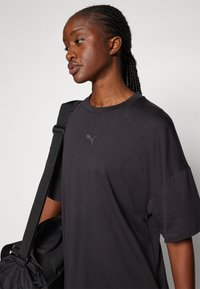 Svart oversized t-skjorte med rund hals, korte ermer og en reflekterende Puma-logo på brystet, kombinert med en svart skulderveske.