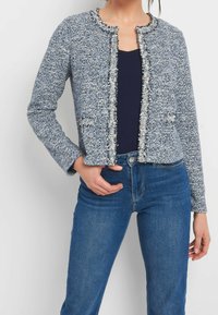 Blå och vit tweedjacka med texturerad yta, kortad design, rund halsringning och fransade detaljer, bärs över en marinblå topp och jeans.