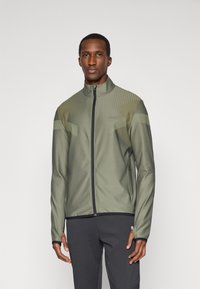 Ελιές πράσινη αθλητική ζακέτα με ψηλό γιακά, κλείσιμο με φερμουάρ, σιλικόνη διακοσμητικά και οριζόντιες ρίγες στους ώμους, συνδυασμένη με σκούρα παντελόνια.