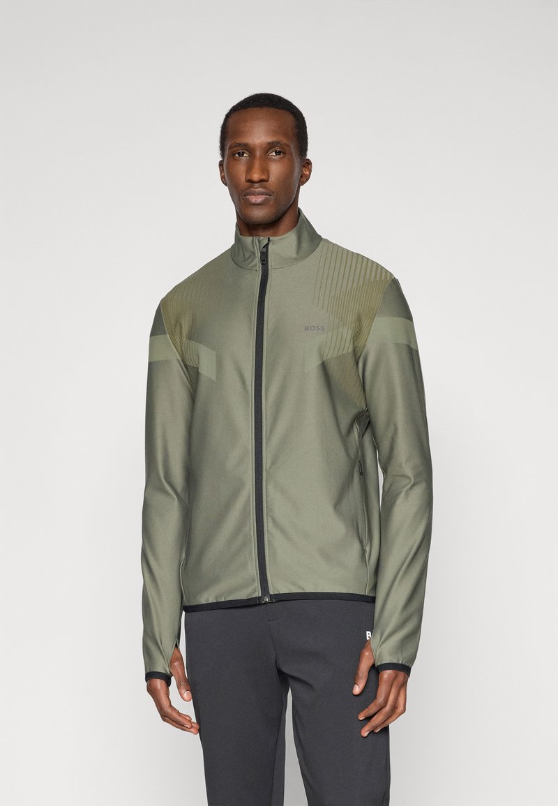 Ελιές πράσινη αθλητική ζακέτα με ψηλό γιακά, κλείσιμο με φερμουάρ, σιλικόνη διακοσμητικά και οριζόντιες ρίγες στους ώμους, συνδυασμένη με σκούρα παντελόνια.