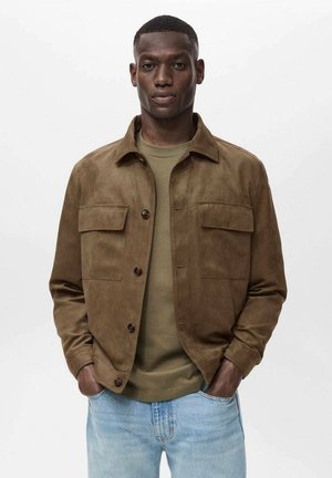 Braune Wildlederjacke mit Knopfverschluss und zwei Fronttaschen, getragen über einem grünen Hemd, kombiniert mit hellblauen Jeans.