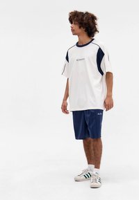 Biele a námornícke športové tričko s krátkymi rukávmi, doplnené o námornícke pruhované šortky. Obsahuje logo a štýlové bežecké tenisky s pruhmi.