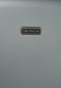 Etiqueta rectangular de metal de la marca ITACA centrada en una superficie de tela de color gris plateado texturizada, probablemente parte del exterior de un equipaje o bolsa.