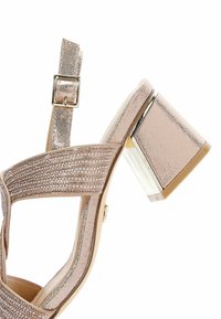 Sandale à talons ouverts en métal rose doré avec un cuir synthétique brillant, des embellissements en cristal sur les brides et un talon bloc transparent.