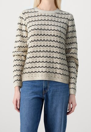 Kvinde iført en cremefarvet strikket sweater med sorte horisontale blonderstriber og blå jeans, stående mod en ensfarvet baggrund.