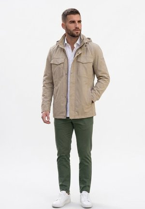 Hombre con chaqueta con capucha beige, camisa blanca, pantalones verdes y zapatillas blancas, de pie con una mano en el bolsillo, mirando hacia un lado.