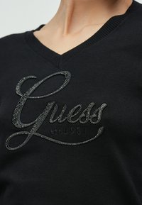 Jersey negro de cuello en V con logo "Guess" en negro brillante con detalles de pedrería y fecha de fundación. Textura suave de la tela.