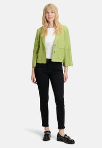 Kobieta z długimi blond włosami, ubrana w jasnoszary krótki żakiet, biały t-shirt, czarne obcisłe dżinsy i czarne loafersy na białym tle.