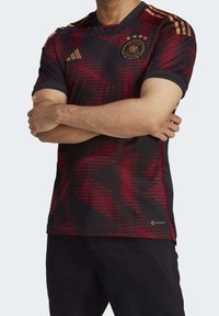 Punainen ja musta jalkapallopaita v-kauluksella, jossa on geometrinen kuvio, kultaiset yksityiskohdat ja joukkueen logo neljällä tähdellä.