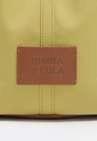 Żółta torba z materiału z brązową skórzaną metką z napisem "BIMBA Y LOLA." Posiada przeszycia i prostokątny kształt. Gładka struktura.