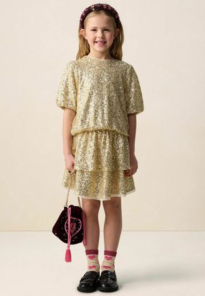 Jeune fille portant une robe en sequins dorés, des chaussettes à motifs de cœurs, des chaussures noires, un serre-tête rose, et tenant un sac à main marron foncé avec un motif en forme de cœur.