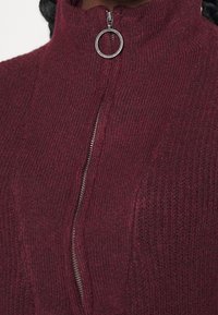 Acercamiento de un suéter de punto marrón con tirador de cremallera en forma de anillo plateado y cuello alto, destacando la textura de la tela y los detalles de la cremallera.