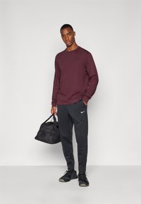 Bordeaux langermet skjorte, svarte joggebukser med hvit Nike-logo, og svart mesh treningsbag. Modellen har på seg svarte treningssko.