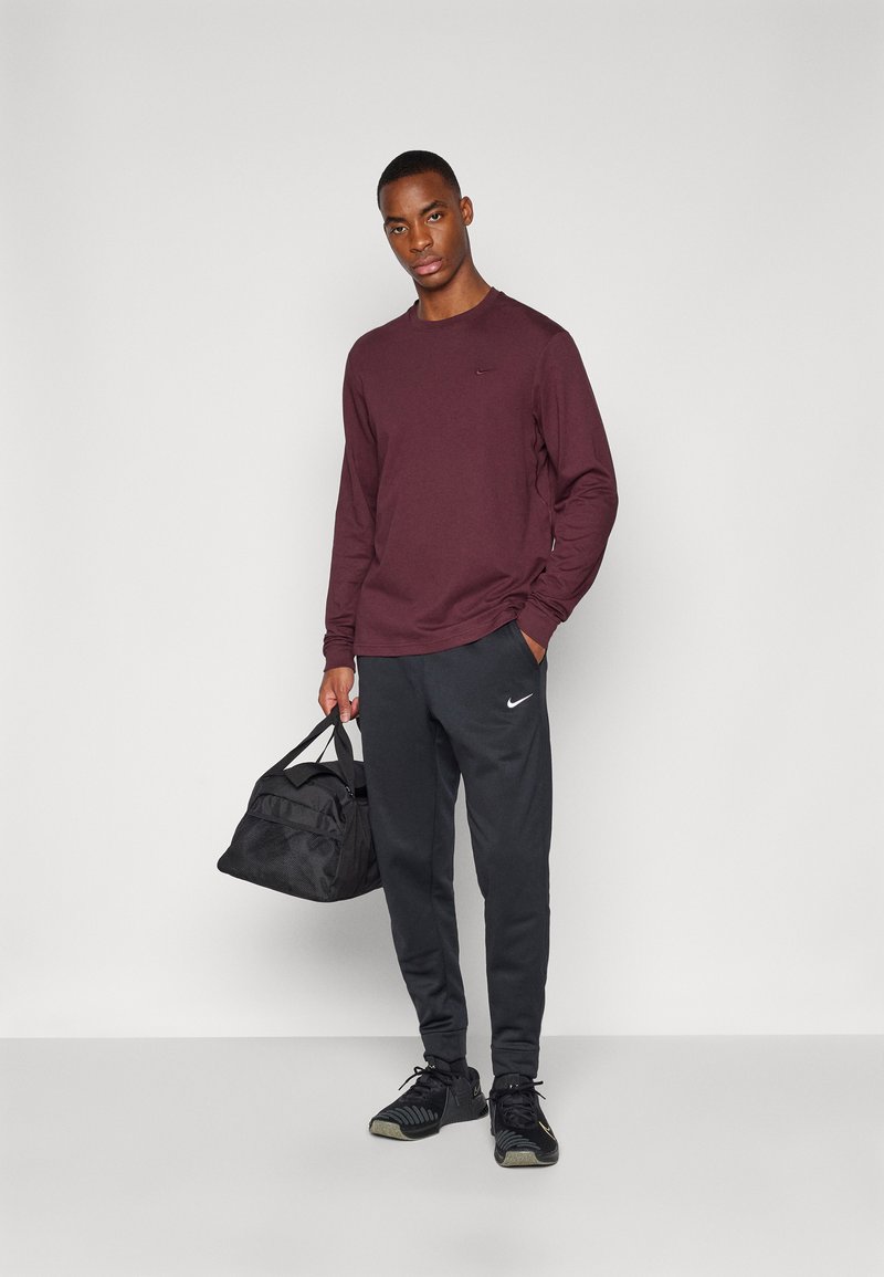 Bordowa koszula z długim rękawem, czarne dresy z białym logo Nike oraz czarna torba sportowa z siateczki. Model ma na sobie czarne buty sportowe.