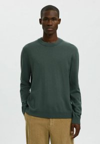 Hombre vistiendo un suéter de cuello redondo verde oscuro y pantalones de pana color beige, de pie con las manos parcialmente en los bolsillos frente a un fondo plano.
