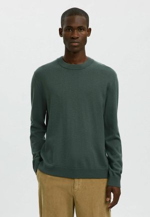 Man in een donkergroen sweatshirt met ronde hals en een lichtbruine corduroy broek, staand met de handen deels in de zakken tegen een effen achtergrond.