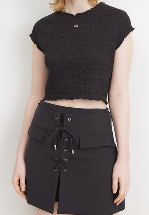 Gros plan d'une personne portant un haut court noir texturé et une jupe noire avec un laçage à l'avant et deux poches à rabat.