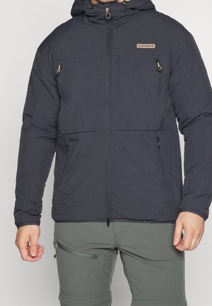 Man wearing a black zip-up hooded jacket with chest and side zippered pockets, paired with olive green pants with a zippered thigh pocket.