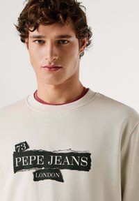 Felpa crema con stampa grafica nera con "PEPE JEANS LONDON" e effetto strappato. Collo a costine, texture morbida. Maglietta interna visibile.