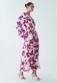 Kreemjas maxi kleit suure purpurse lillemustriga, sügava V-kaelusega, volangiliste varrukatega ja korsetiümbrisega taljeosas. Kandke punutud kontsaga sandaalidega.