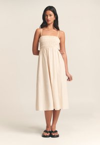 Vaaleanbeige midi-mekko, jossa on rennosti rypytetty yläosa, ohuet olkaimet ja hulmuava helma. Yhdistetty mustiin tasapohjaisiin sandaaleihin, joissa on yksinkertainen ja minimalistinen muotoilu.