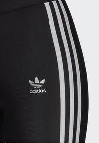 Mustat urheilulegginsit, joissa on kolme pystysuuntaista hopeista ADDI-kangasta sivuilla. Vasemmassa reidessä on pieni valkoinen Adidas-logo.