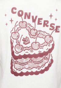 Λευκό μπλουζάκι με γραφικό σε μαρόν χρώμα που απεικονίζει μια τούρτα με στρώσεις, διακοσμημένη με κεράσια και μια μικρή γάτα στην κορυφή, καθώς και διακοσμητικά αστέρια και περιγράμματα.