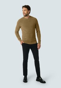 Bruine, gestructureerde gebreide trui met een ronde hals, gecombineerd met zwarte slim-fit broek en gepolijste zwarte schoenen, tegen een lichte achtergrond.