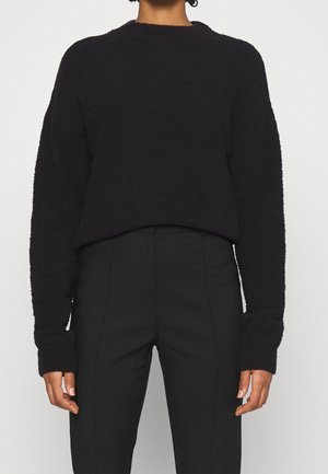 Osoba nosící černý strukturovaný svetr s dlouhým rukávem a černé vysoké kalhoty na míru na jednobarevném bílém pozadí.