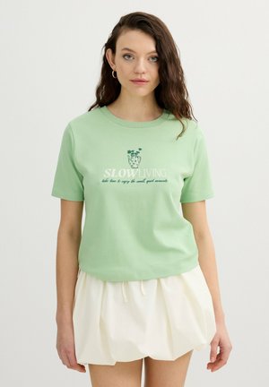 Mujer con cabello castaño ondulado, que lleva una camiseta verde claro con el texto "SLOW LIVING" y una falda blanca suelta sobre un fondo claro y liso.