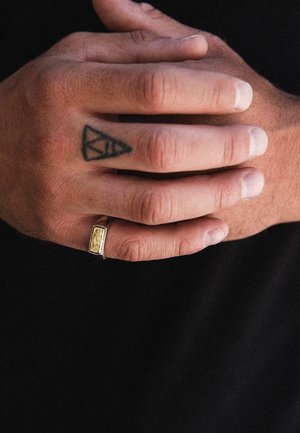 Gold-toned rectangular ring with textured surface, worn on a hand with a tattoo, fingers slightly curled, showcasing skin tone and nail color.