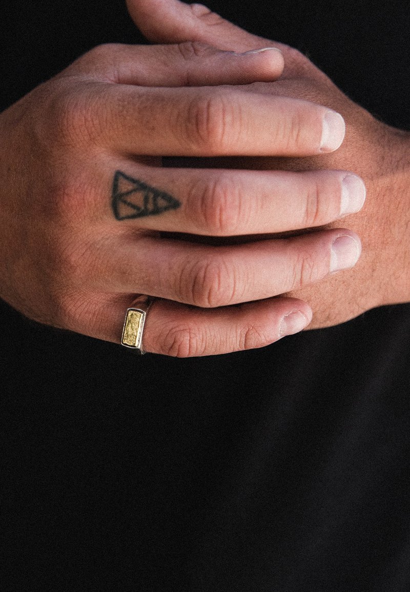 Auksinės spalvos stačiakampio formos žiedas su tekstūruotu paviršiumi, dėvimas rankoje su tatuiruote, pirštai šiek tiek sulenkti, demonstruojantys odos atspalvį ir nagų spalvą.