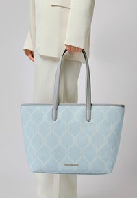 Persona in un completo crema tiene in mano una borsa a spalla leggermente blu con motivo, dotata di manici grigi e di un piccolo logo dorato del marchio.