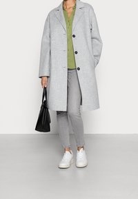 Cappotto grigio di lana con bottoni neri, camicia verde sotto, jeans grigi e sneakers bianche. Borsa nera in mano. Texture lisce e design semplice.