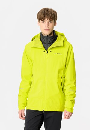 Jeune homme portant une veste imperméable Vaude jaune vif avec capuche et un pantalon noir, debout devant un fond clair uni.