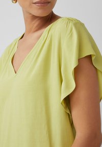 Lichtgroene blouse met een V-hals en fladdermouwen. Zachte textuur en subtiele glans versterken het vloeiende ontwerp.