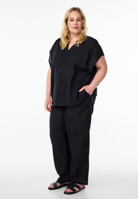 Musta pellavapaita ja leveälahkeiset housut, joissa V-pääntie, lyhyet hihat ja sivutaskut; yhdistetty mustiin, niitein varustettuihin sandaaleihin.
