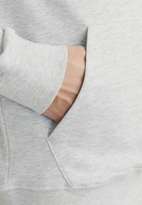 Sudadera gris claro con puños acanalados y un bolsillo canguro en la parte delantera, que muestra una tela suave y texturizada y un detalle de costura oculta.