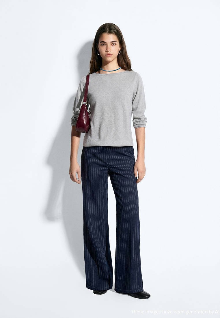 Jeune femme debout portant un haut à manches longues gris clair, un pantalon large bleu marine à fines rayures, des chaussures noires, portant un sac à épaule bordeaux.
