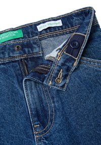 Modré džínové kalhoty s částečně rozepnutým zipem, knoflík na pase zapnutý a viditelné zelené a bílé značkové štítky.