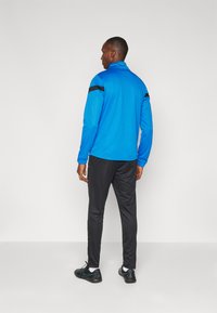 Veste de sport bleue avec un col haut, accents noirs sur les épaules, associée à un pantalon de survêtement noir et des chaussures de sport noires. Texture lisse.