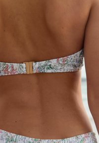 Bikiny top v bílé látce s vícebarevným květinovým vzorem, s hladkým, naříznutým pásem a zlatým zapínáním na zádech.