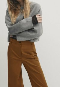 Pull gris surdimensionné à manches larges associé à un pantalon marron taille haute avec une fermeture à un bouton et une texture lisse.