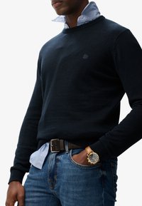 Marineblå sweater med rund hals, båret over en lyseblå, skjortekræset skjorte. Mørke jeans og et guldur med en brun læderrem fuldender outfittet.