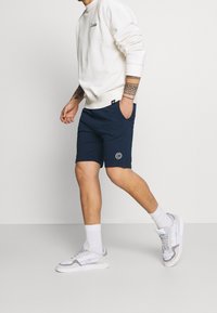 Laivastonsiniset shortsit, joissa on taskut ja pyöreä logo, yhdistetty vaaleaan kermaiseen collegepaitaan ja valkoisiin tennareihin, joissa on harmaita yksityiskohtia.