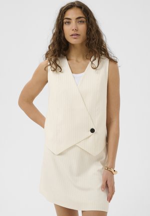 Jonge vrouw met krullend haar, gekleed in een mouwloze crèmekleurige pinstripe vestjurk en een gouden armband, staand tegen een effen achtergrond.