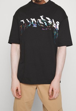 Person wearing a black oversized T-shirt with distorted multicolor text "TIMELESS" across the chest and beige pants against a plain background.